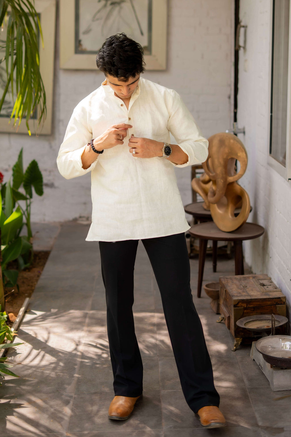 Man wearing an off-white linen short kurta with a mandarin collar, paired with black trousers and brown shoes, standing in a corridor with plants and artistic wooden decor.