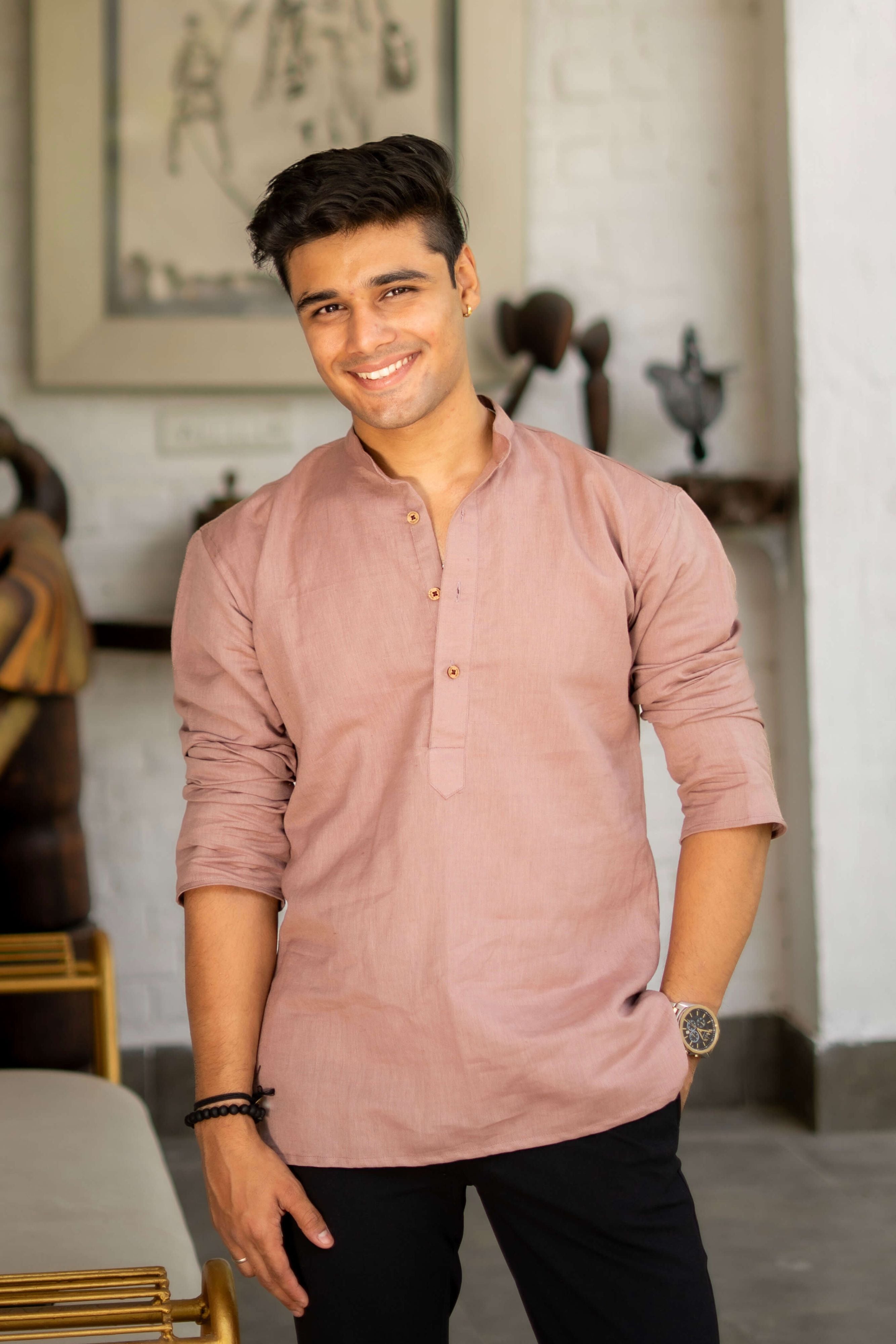 Man smiling and wearing a light purple linen short kurta with wooden buttons, standing indoors with natural sunlight and green plants in the background.