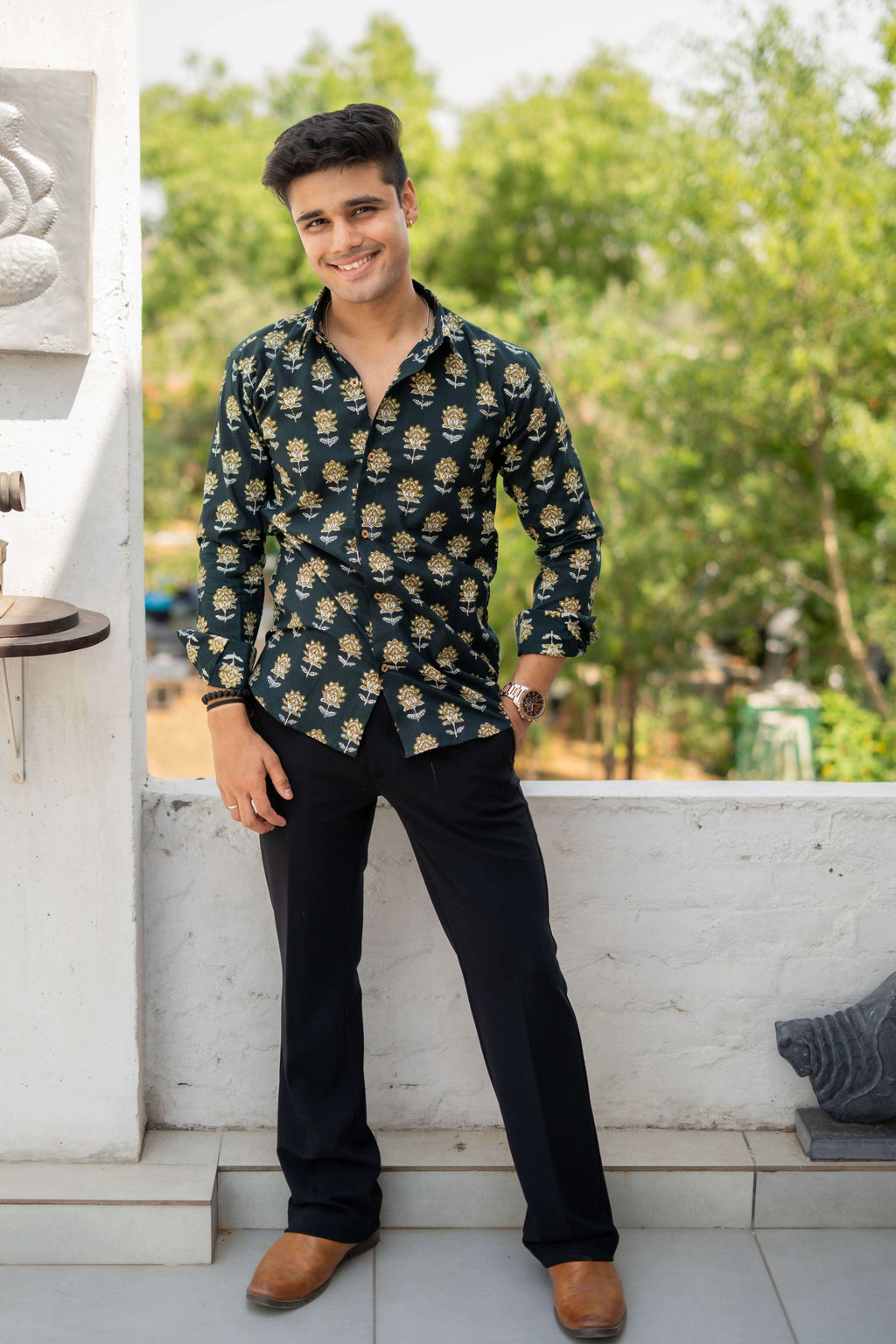 An Indian man wearing a Castel green shirt with a yellow and white flower print, styled casually in an outdoor setting. He pairs it with a black beaded bracelet and a watch, surrounded by greenery.