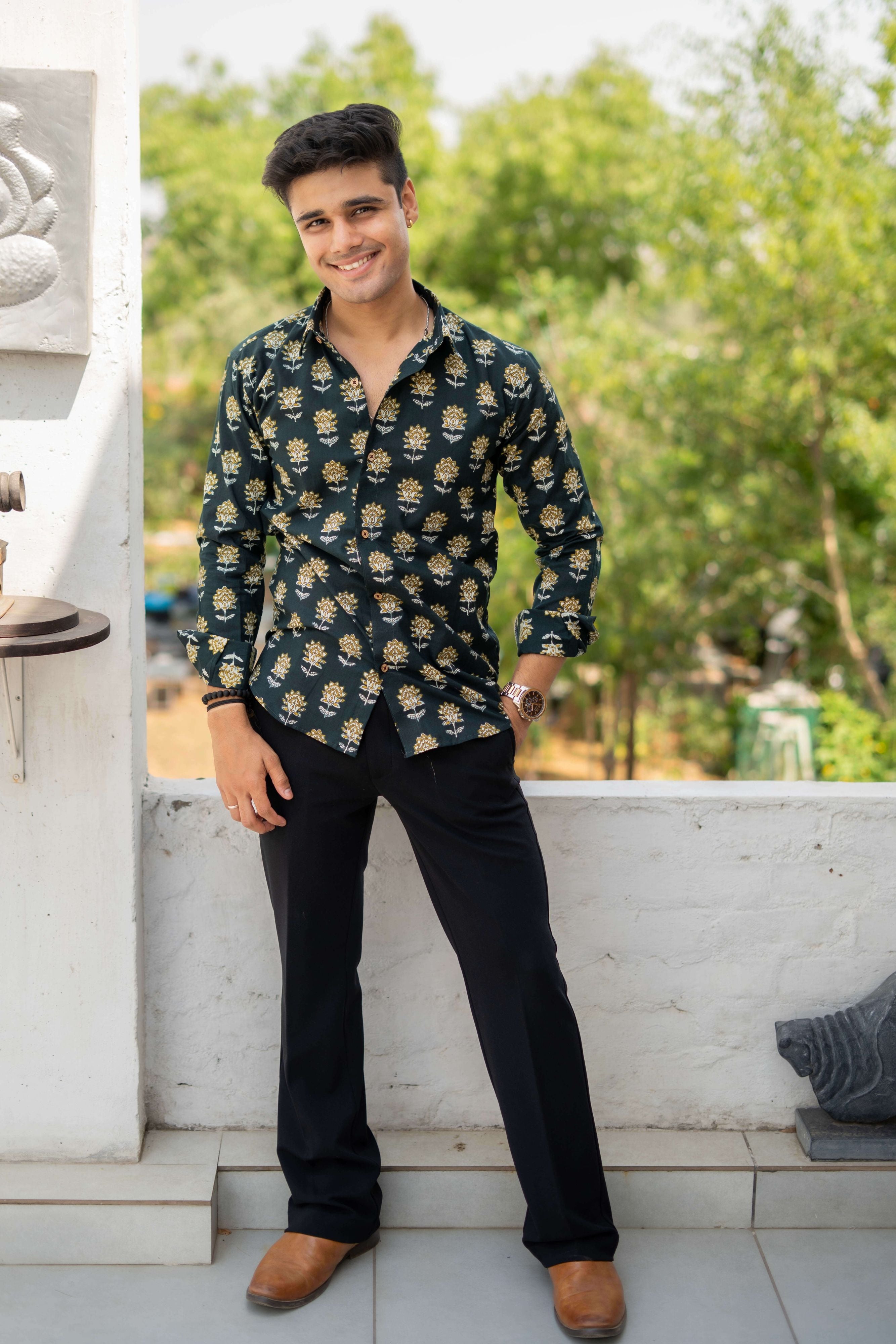 An Indian man wearing a Castel green shirt with a yellow and white flower print, styled casually in an outdoor setting. He pairs it with a black beaded bracelet and a watch, surrounded by greenery.