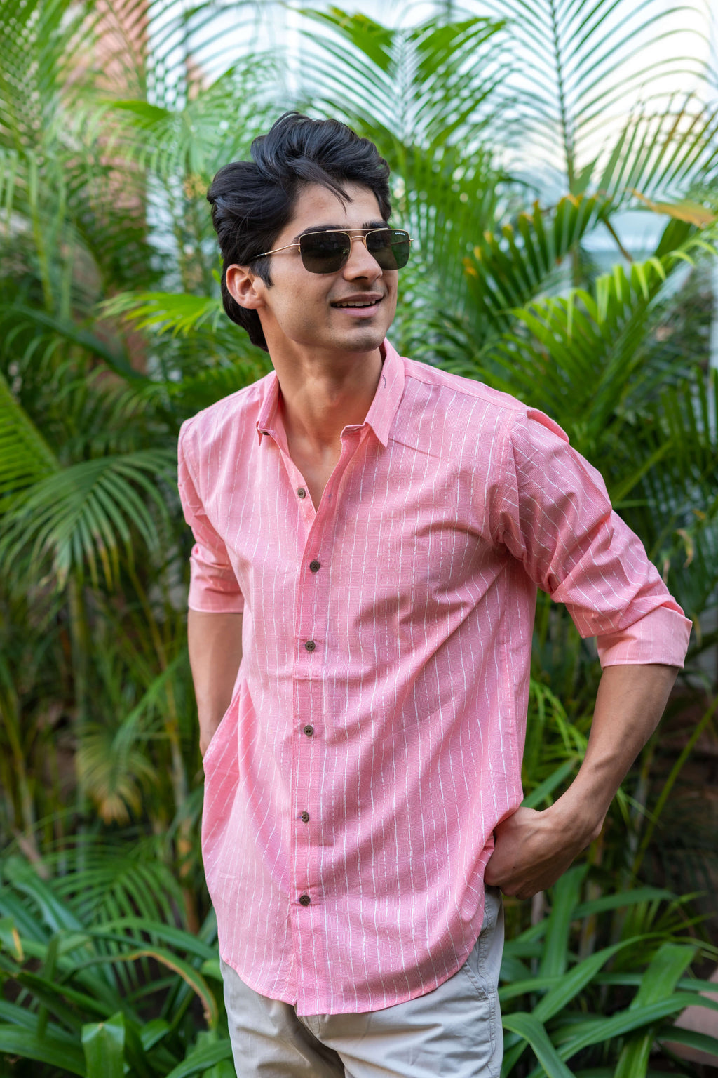 An Indian Man in a Pink Striped Shirt and shorts Is Standing in Front of Green Palm Leaves, Wearing Sunglasses and Smiling 