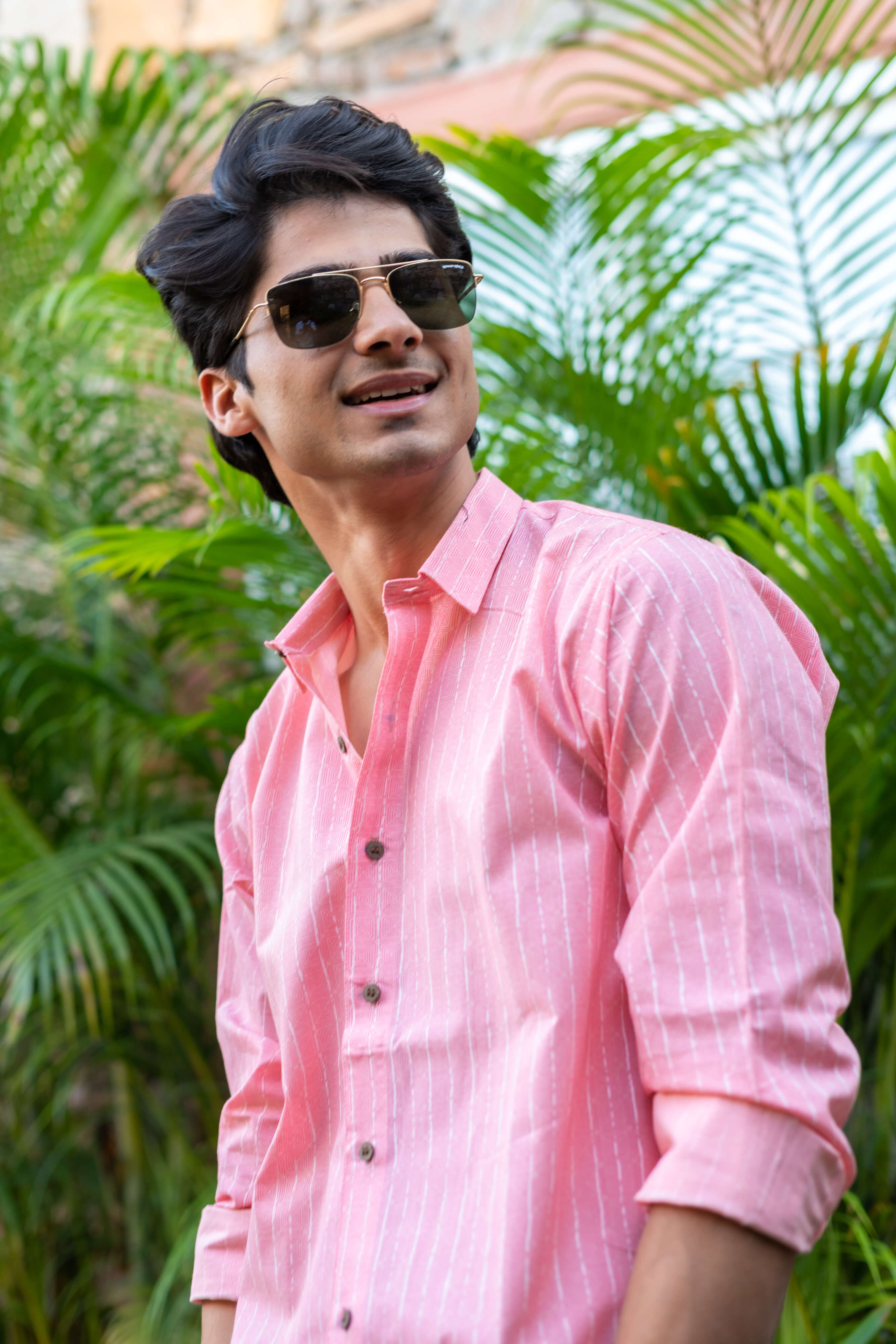 An Indian Man in a Pink Striped Shirt Is Standing in Front of Green Palm Leaves, Wearing Sunglasses and Smiling posing for side angle