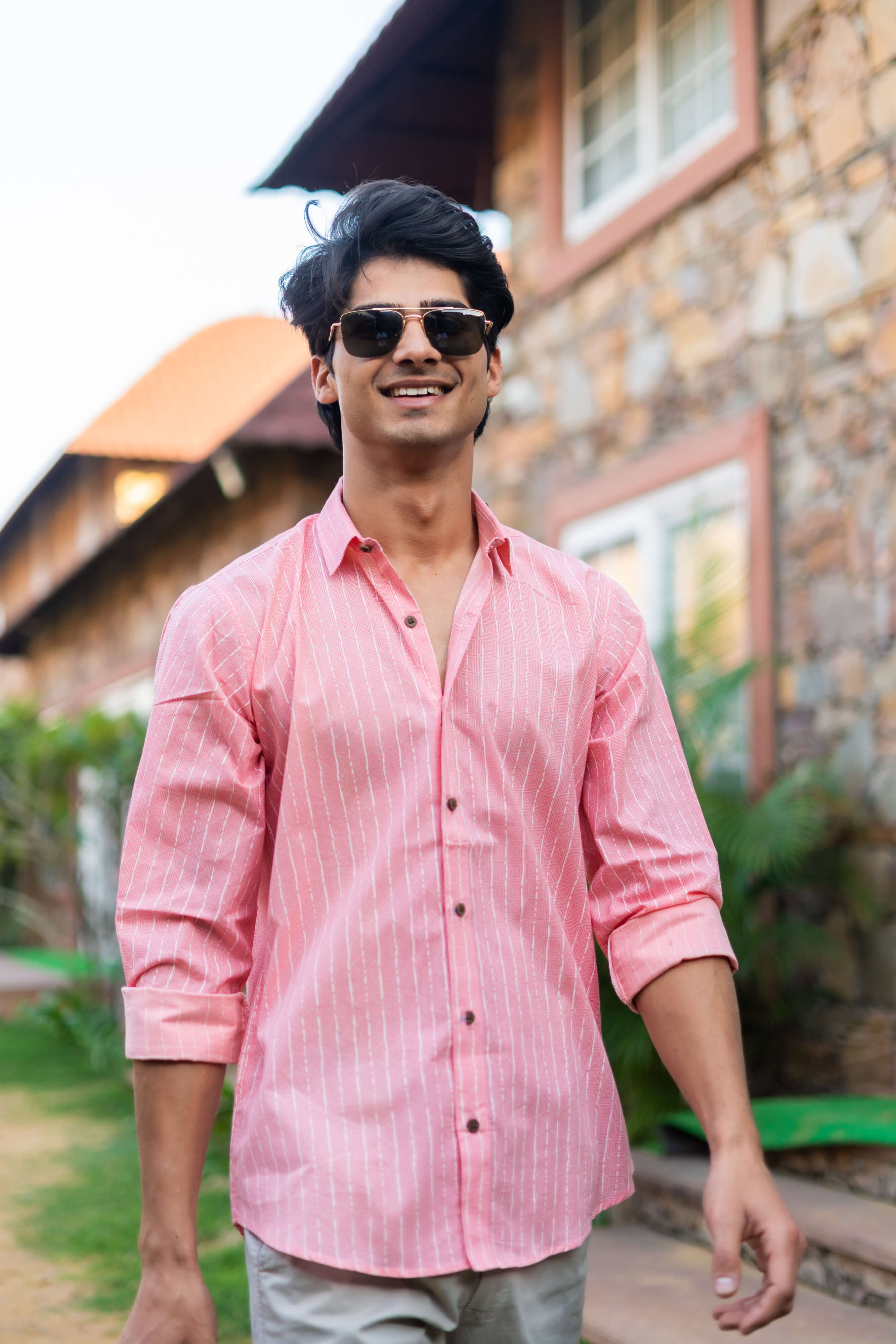 An Indian Man in a Pink Striped Shirt Is Standing in Front of a wall, Wearing Sunglasses and Smiling.