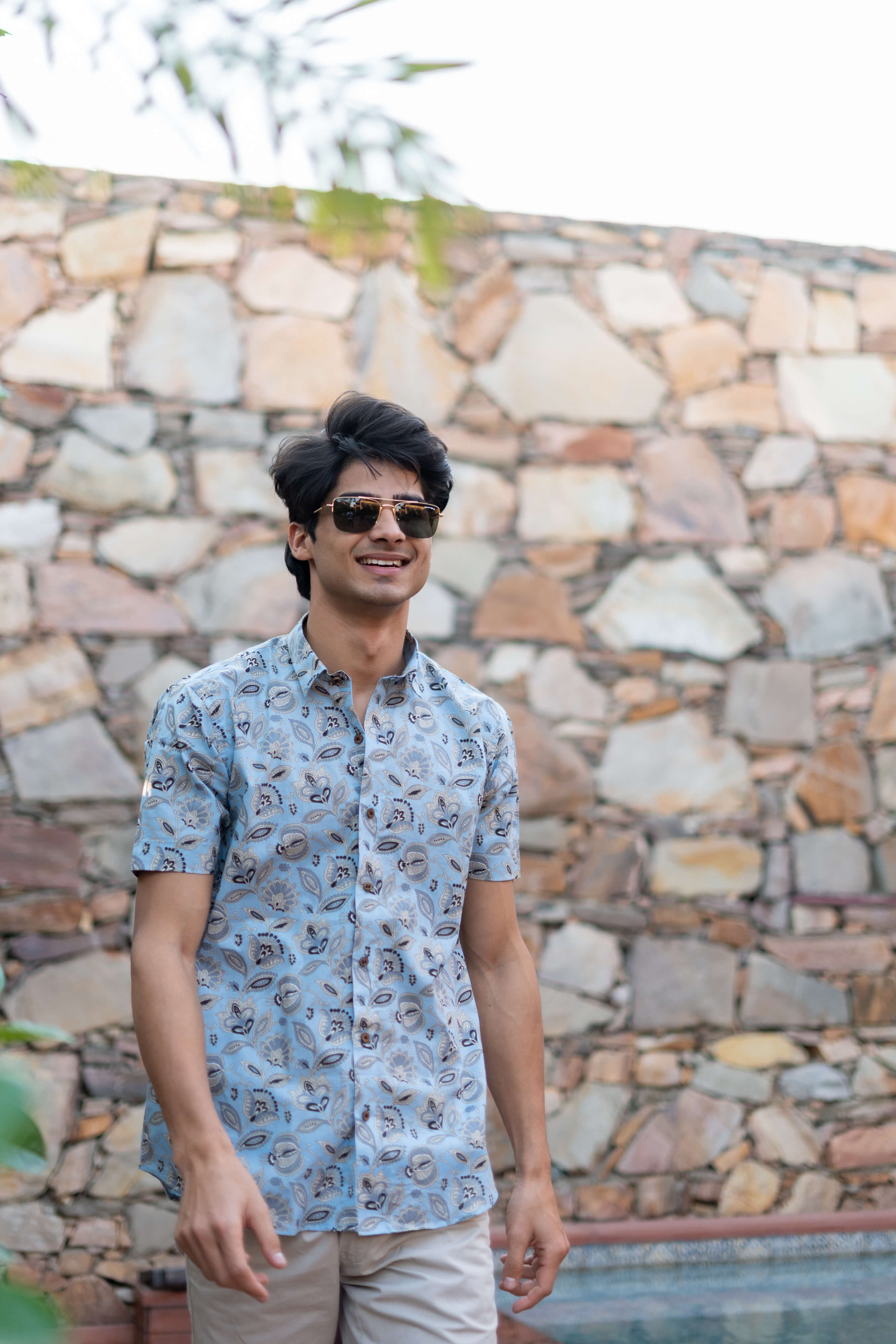 An Indian Man Wearing a Funky Sky Blue Color Half Sleeves Shirt with a Floral Print and Sunglasses, Outside in Front of a Stone Wall.