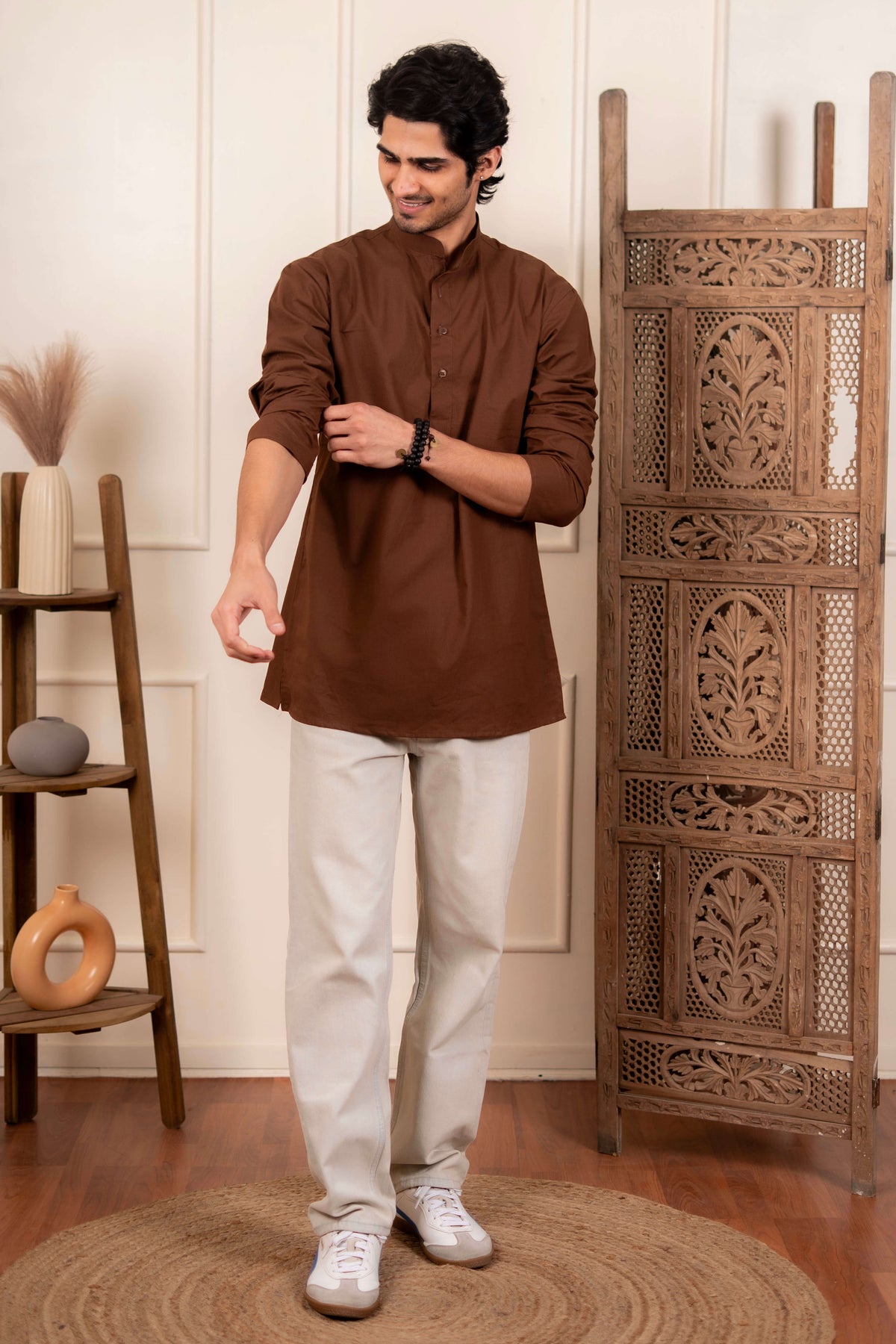 Man wearing a solid brown short kurta with rolled-up sleeves, paired with off-white pants and white sneakers, standing indoors against a decorative wooden panel and minimal home decor.