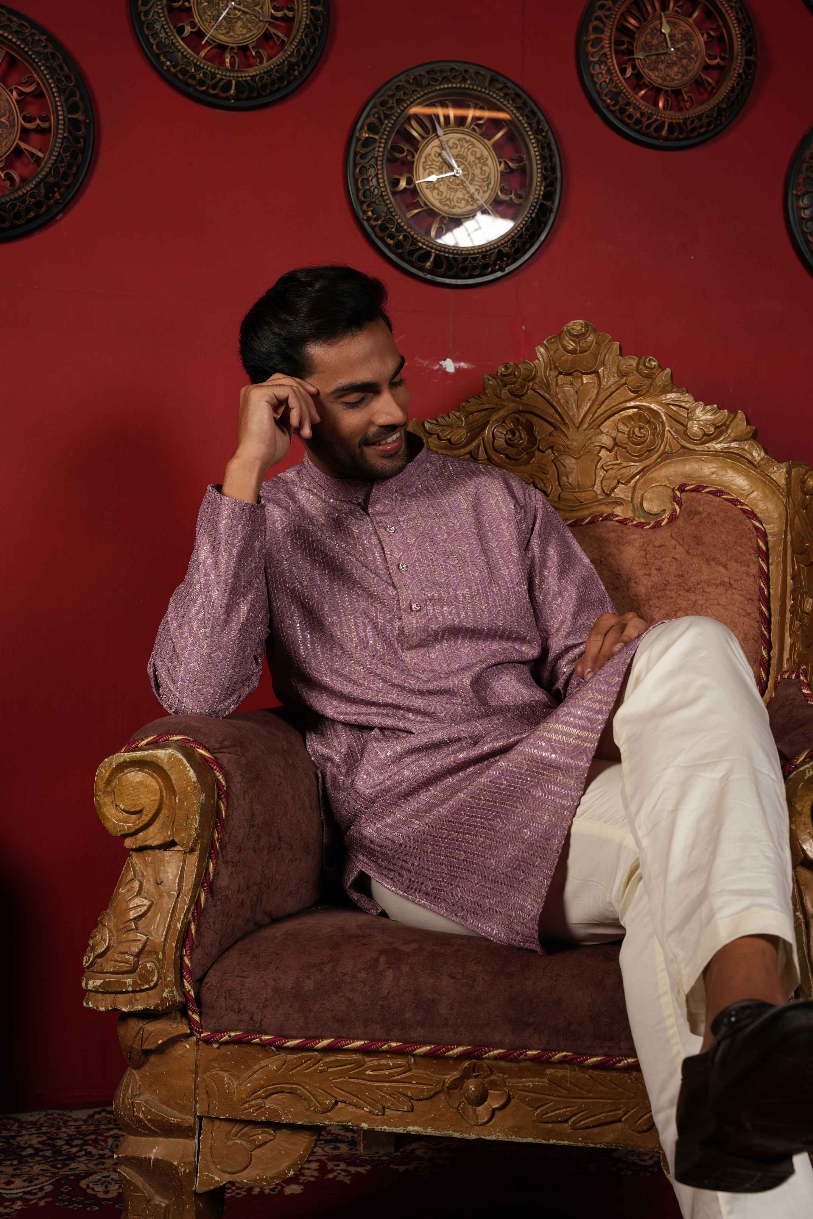 Man in a purple traditional outfit sitting on an ornate chair against a red wall with decorative clocks.