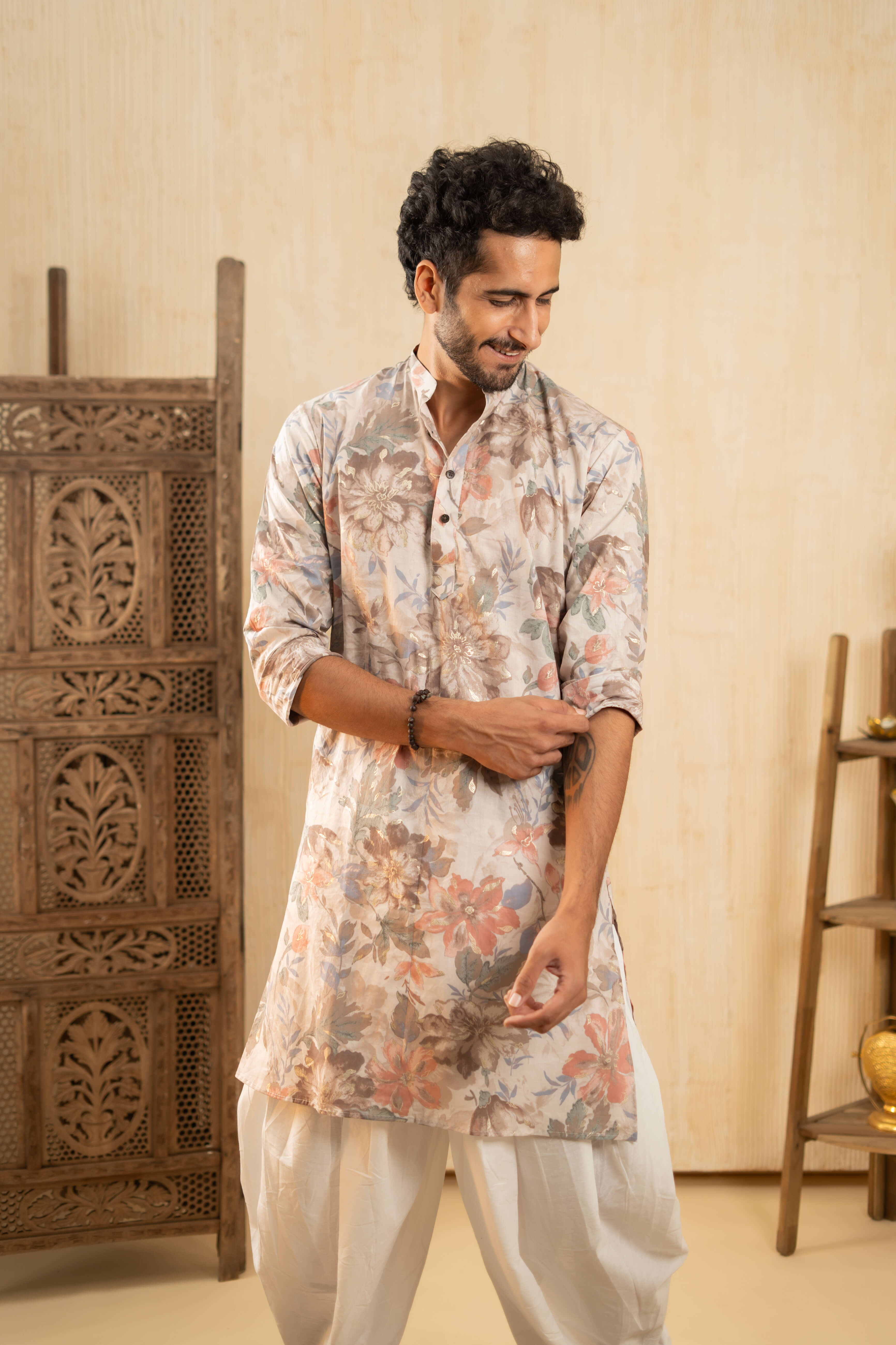 Indian man wearing a floral printed long kurta and white dhoti, posed in an elegantly decorated room.