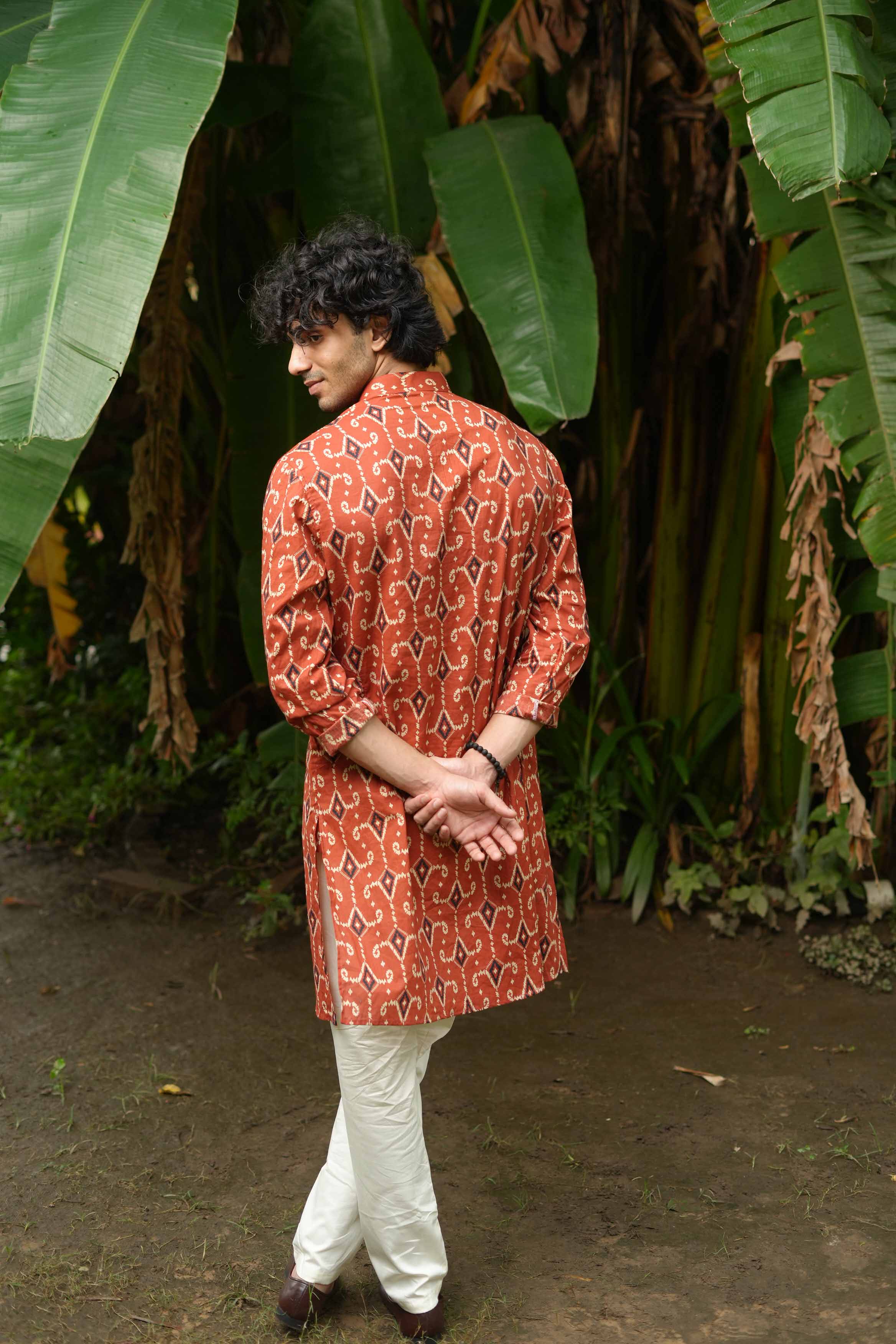 Person wearing a red patterned kurta with white pants standing in front of large green leaves.